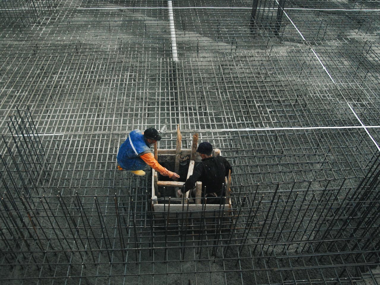 Two workers operating on a construction site with steel frameworks and concrete. High-angle view captures teamwork and industry.