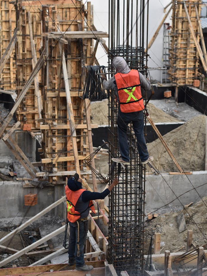 Two workers in safety vests climb a steel structure on a busy construction site.