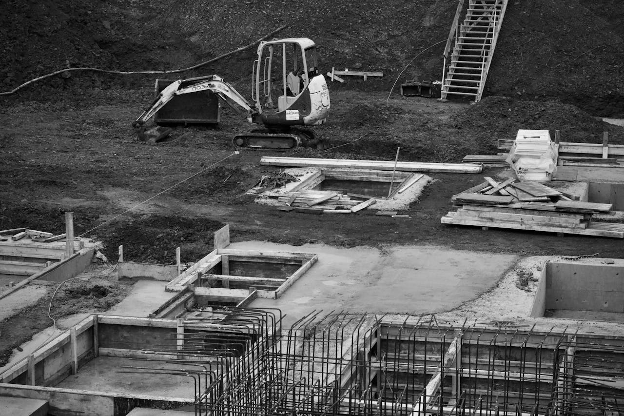 Monochrome image of an excavator at a construction site with foundations laid and rebar in place.